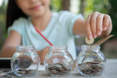 Enfant mettant une pièce de monnaie dans une des trois tirelires transparentes sur une table.