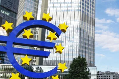 Grand symbole de l'euro devant un bâtiment de banque moderne avec des étoiles de l'Union européenne, sous un ciel bleu.