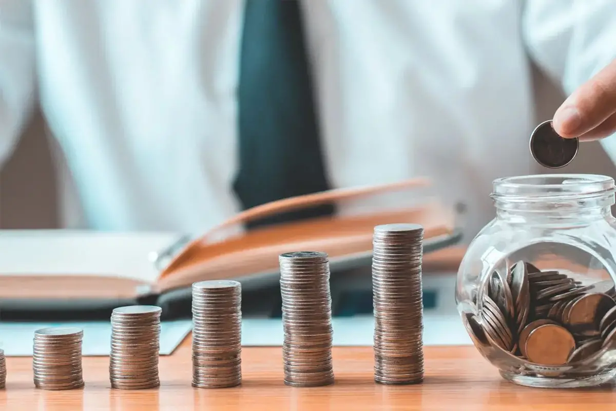 Personne en costume ajoutant une pièce dans un bocal à côté de piles de pièces de monnaie sur un bureau.